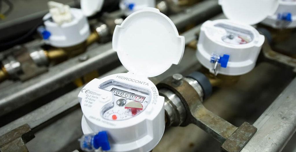 Gros plan mains plombier gantées installant compteur eau numérique résidentiel et clapet anti-retour chromé sur raccordement conduite, outils professionnels visibles, fond mur béton sous-sol